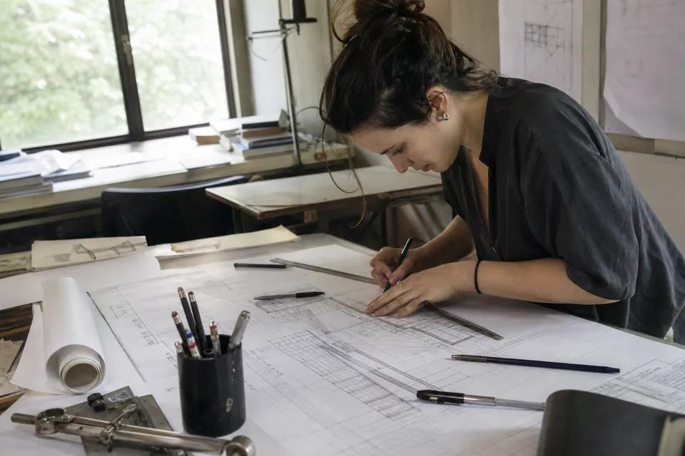 Architect drawing on a drafting table covered with plans, rulers and pencils in daylight