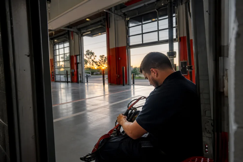Fire station bay with tall doors open at dawn, a firefighter checking gear on the floor