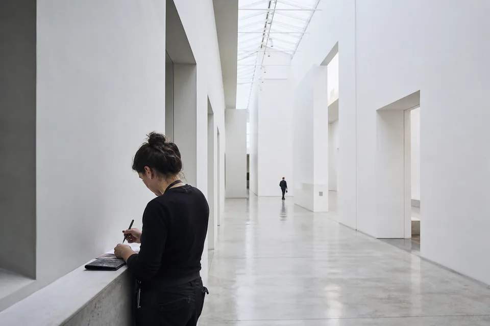 Large white gallery with tall volumes under a skylight, a visitor taking notes
