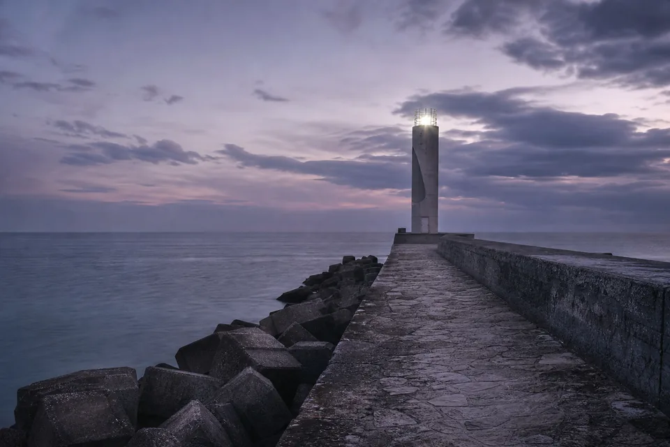 Modern concrete lighthouse on a long breakwater at dusk with its light on