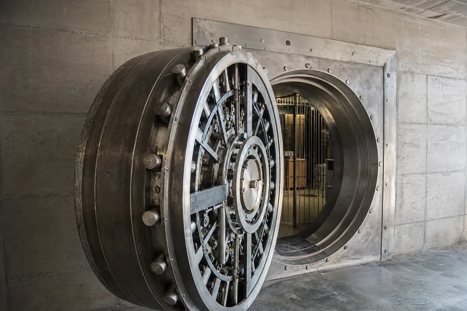 Massive round steel vault door set in a concrete wall, slightly open, lit by daylight